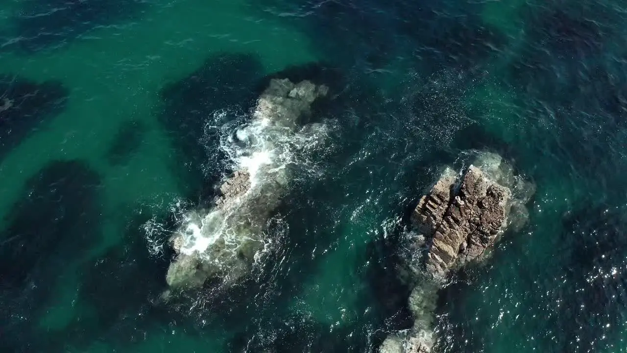 Aerial top down view of ocean waves crashing into small rocks in shallow water