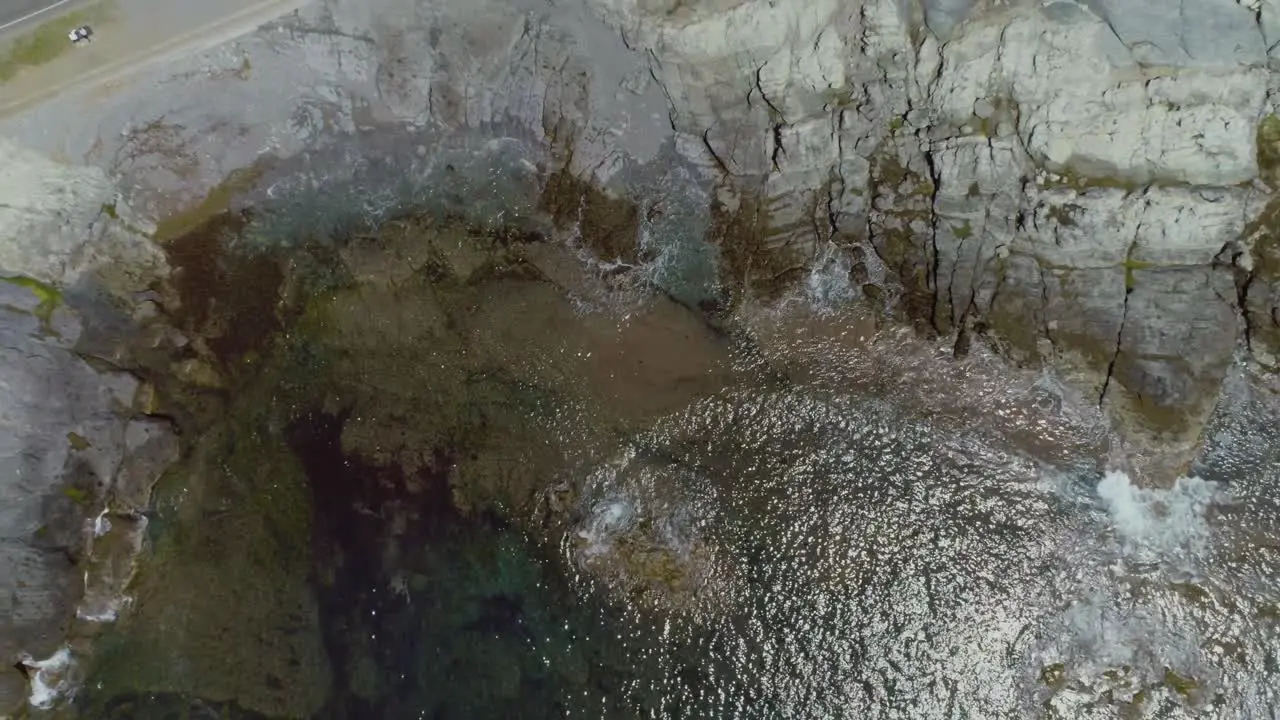 Top-down aerial of tropical ocean water waves crashing on a rocky shoreline during a summer day