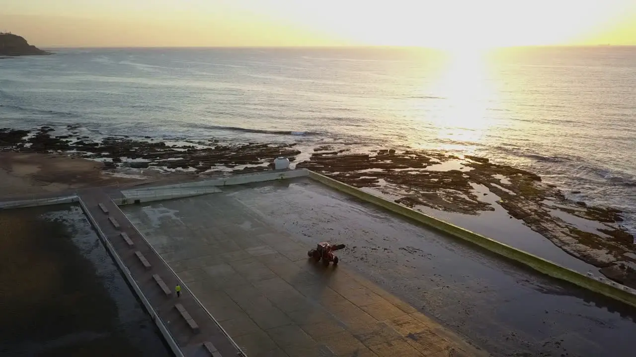 Rotating sunrise aerial Tractor cleans empty beach Mereweather pool