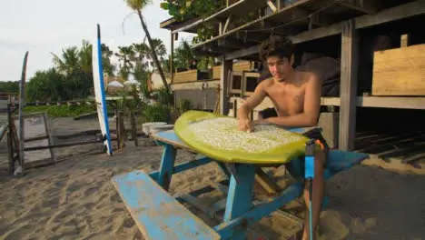 Tracking Shot Pulling Away from Young Man Preparing Surf Board