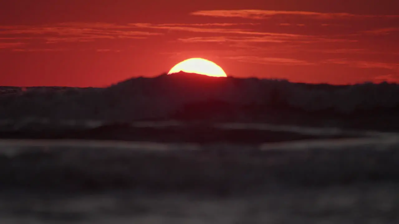 Ocean waves roll across water as yellow ball of sun sets below horizon with red sky telephoto static