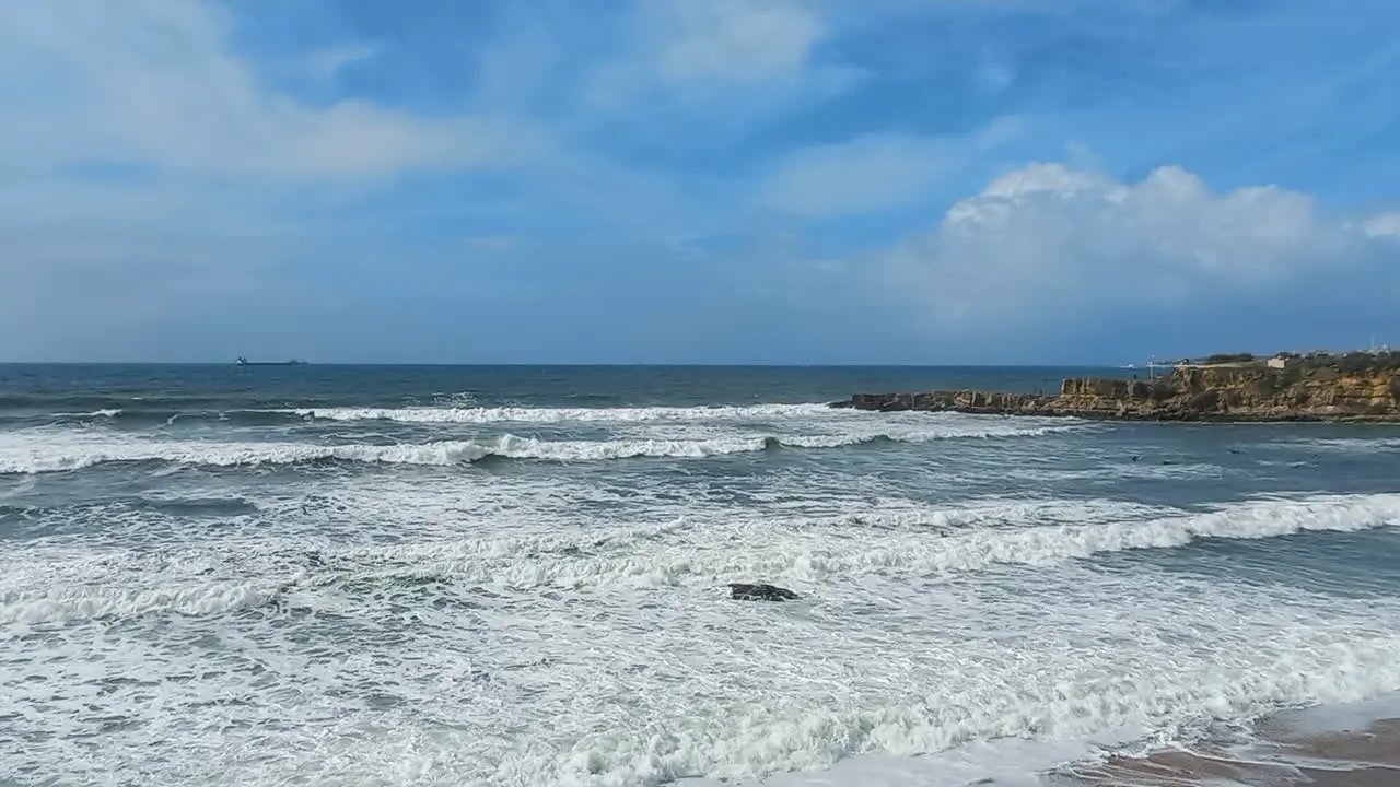 Rough sea creating several waves of white foam some brave surfers and the blue sky with lots of clouds