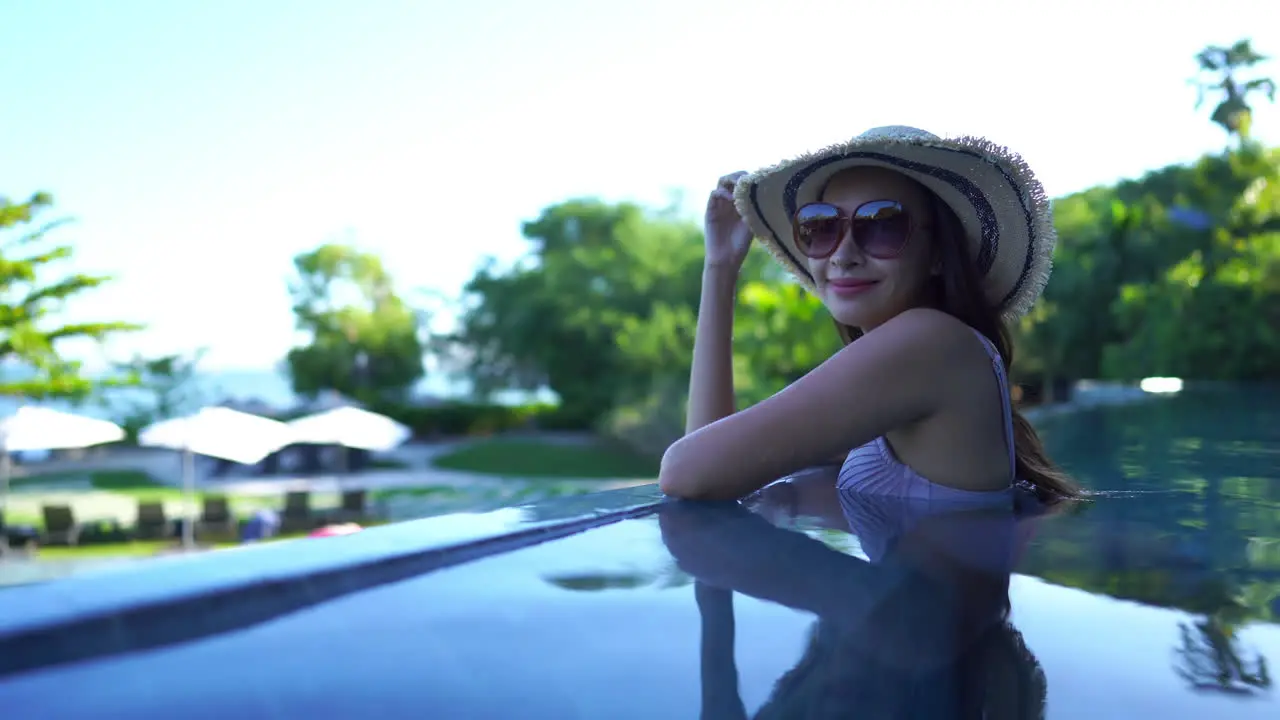 attractive asian woman wearing a striped sunhat and large sunglasses smiling as she enjoys a shady infinity pool with glassy water and bright sunlight on the green trees in the background