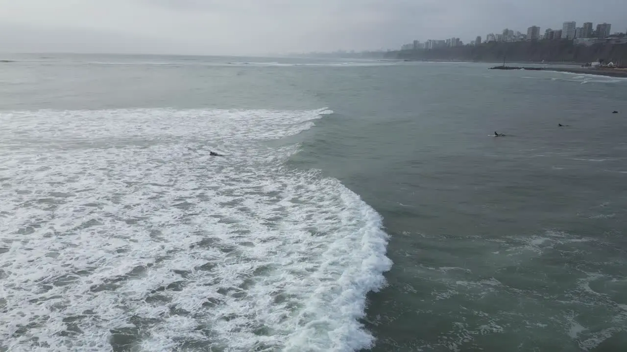 Aerial panning shot of the sea in Lima Peru with calm waves and surfers with surfboards in the water on a cloudy day