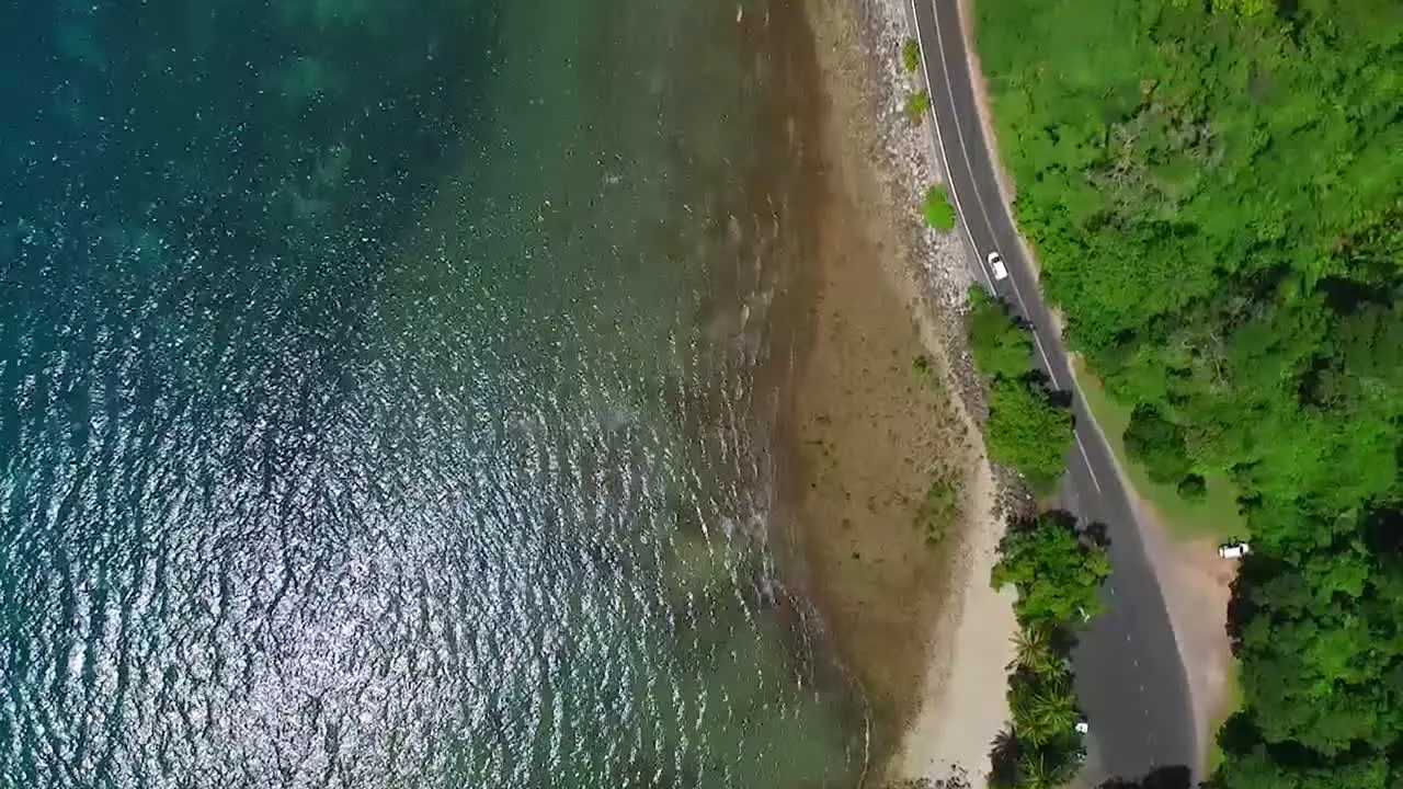 Birdseye view following a white car along a stretch of coastal road on a sunny day