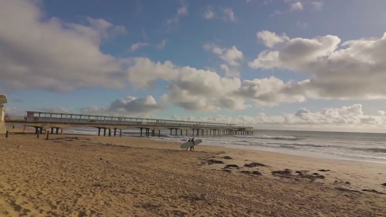 Two surfers with boards walking on the beach towards group of surfers