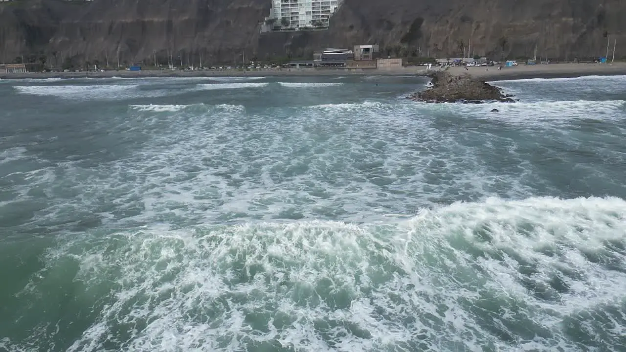 Slow motion aerial dolly shot of the sea with waves in front of the beach of miraflores lima in peru with rocks and building in the background