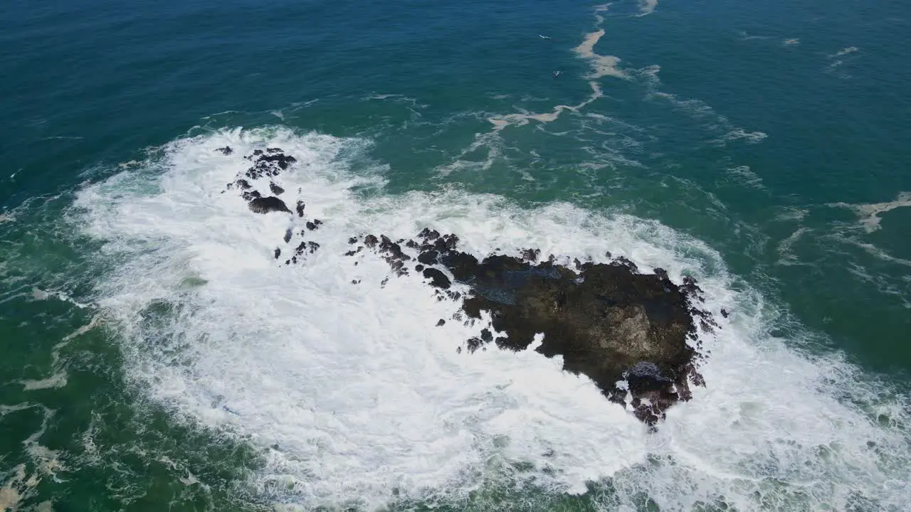 Aerial top down shot of giant waves crashing against coral rock in sunlight