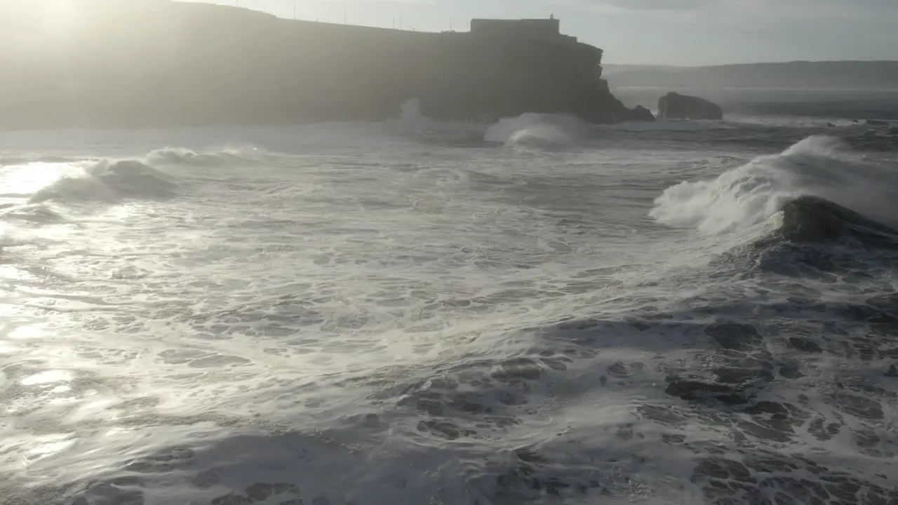 Drone flyover powerful ocean waves breaking toward Nazare Lighthouse promontory sunbeam reflection