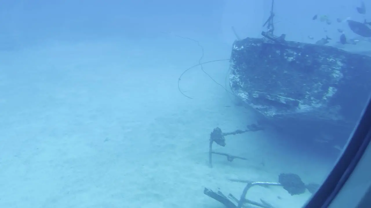 Cinematic wide dolly shot of a fish swimming by a submarine porthole with an eerie shipwreck behind it on the ocean floor off the coast of Hawai'i