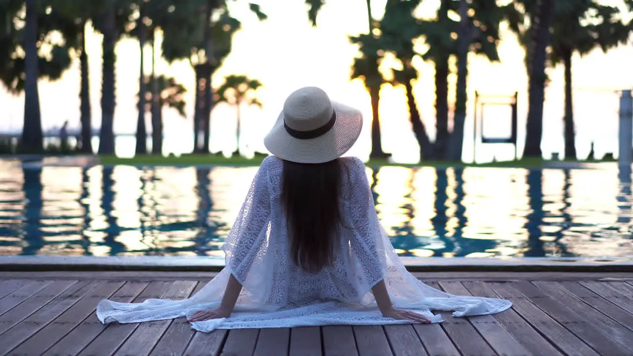 Back of Stylish Female With Summer Hat Sitting on Poolside and Watching Golden Hour Sunlight on Sea Horizon