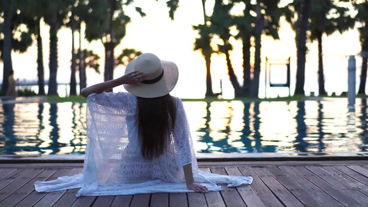 Lonely Stylish Female With Summer Hat and Cloak Sitting By Pool of Luxury Hotel Static Back Shot