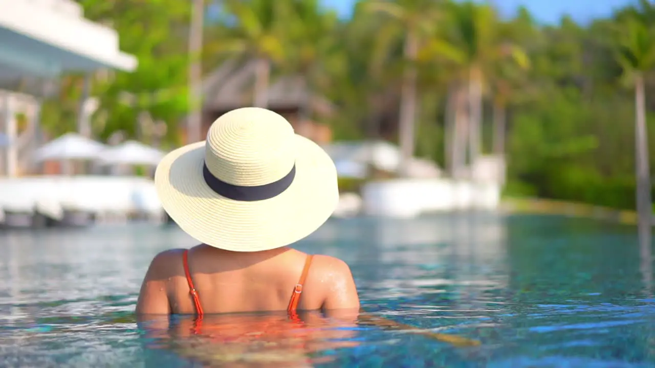 Backside view of an Unrecognizable Pretty Young Model in Swimming Suit and Sunhat Standing Inside the Pool of Tropical Island Resort in Bali Indonesia blurred palm trees on background