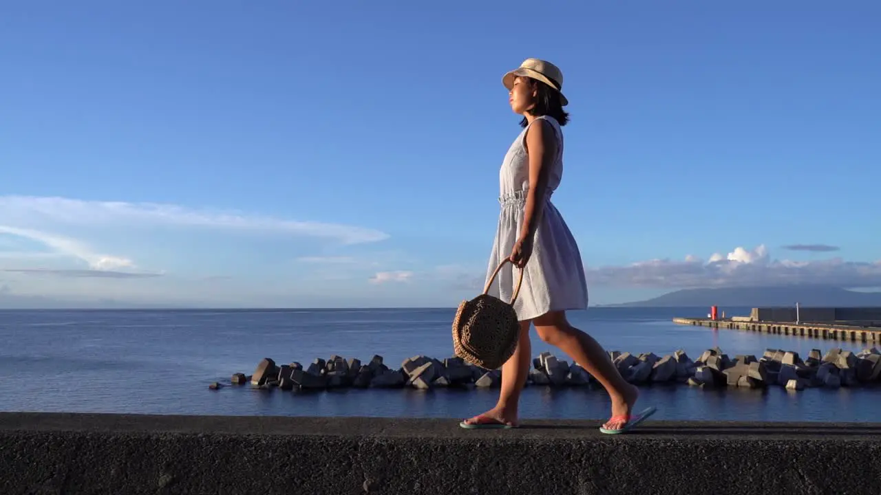 Japanese girl in summer dress holding bag walking in front of Ocean on beautiful clear day wide sideways tracking shot