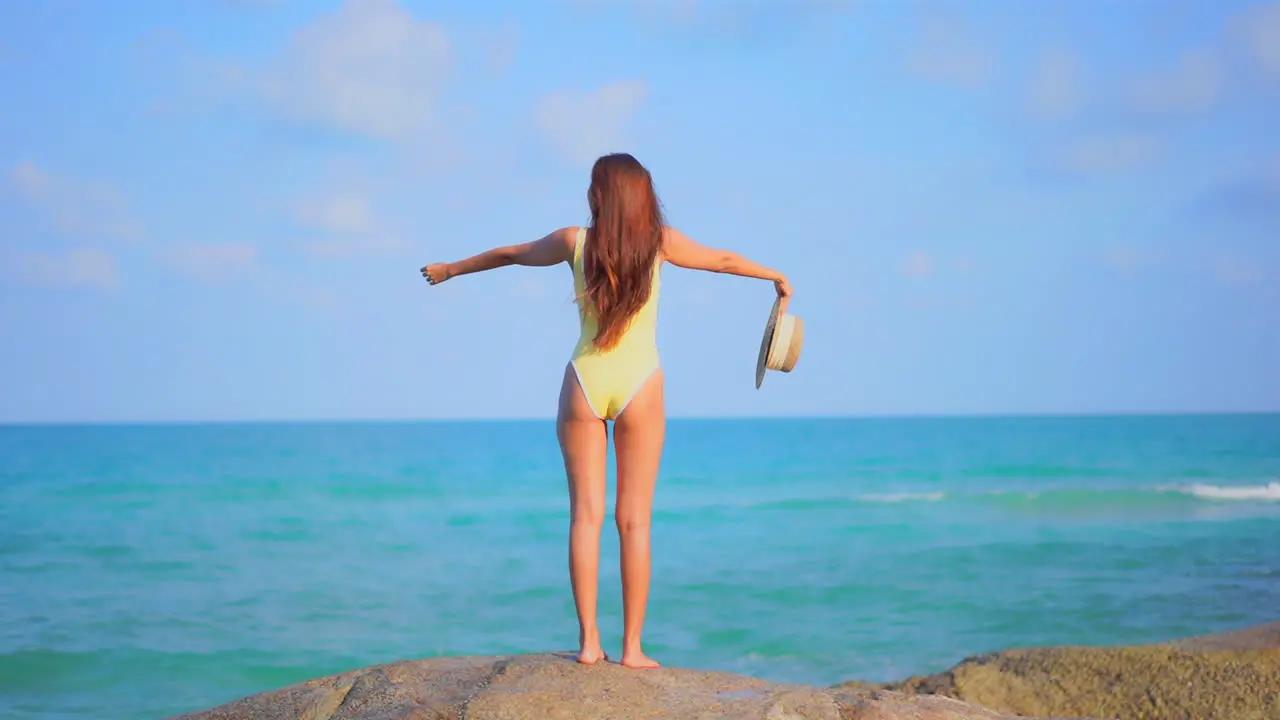 Back to the camera a healthy fit woman in a bathing suit stands on a huge rock facing the ocean horizon