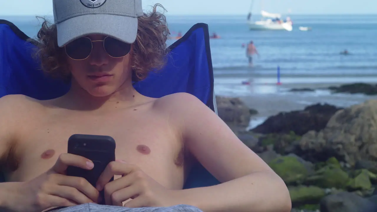 Curly-haired Man Sitting And Using Smartphone At Coverack Beach In Cornwall UK