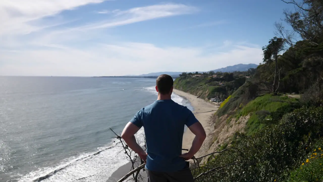 A man standing on the edge of an ocean cliff on a nature hiking trail with an epic beach view on a sunny day in Santa Barbara California SLOW MOTION