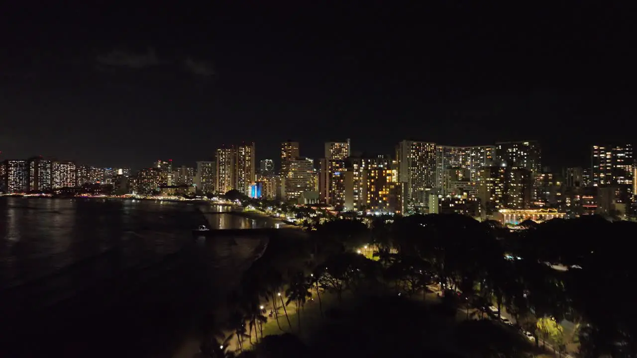 reveal drone aerial night shot of waikiki showing strip and city lights downtown in evening behind trees