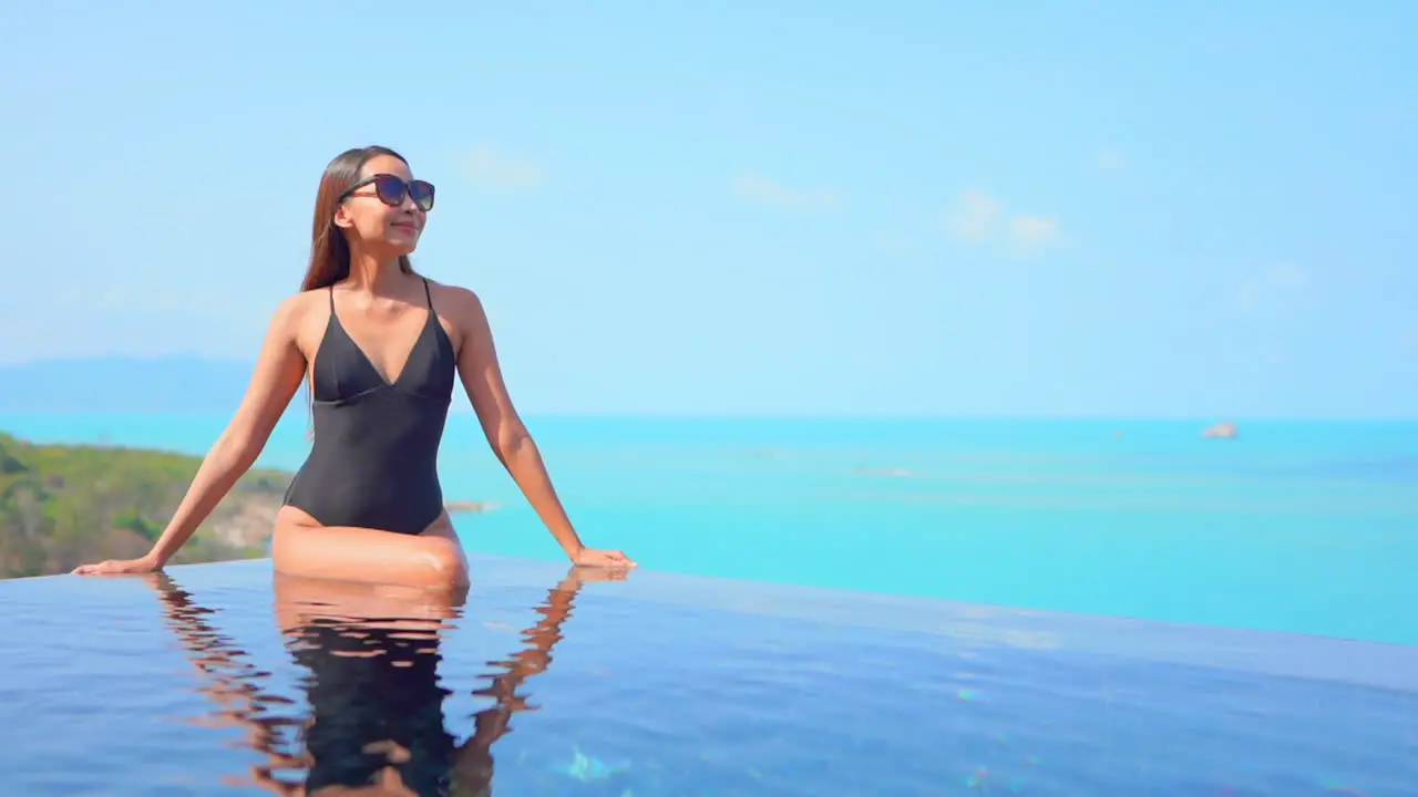 Elegant Asian woman with black swimsuit sit on edge of infinity pool overlooking ocean Slow-motion