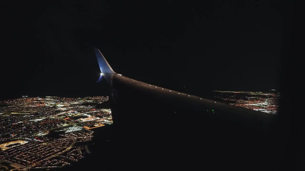 Plane landing in an airport next to a city at night from the inside looking outside at the wing