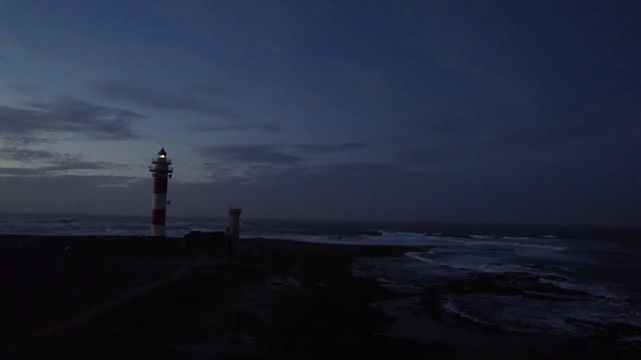 aerial footage of a lonely lighthouse at the rocky ocean shore with yellow light on top at dawn with waves crashing all around and dark blue sky in back