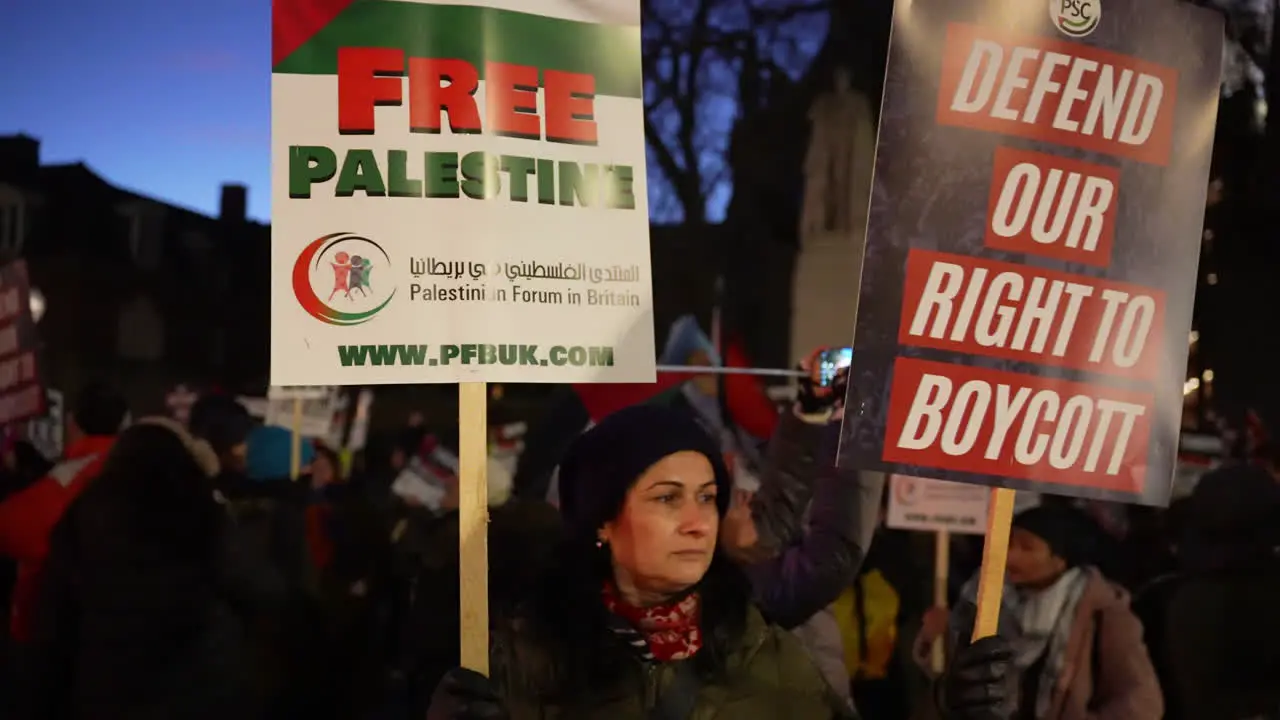 A protestor holds up two placards that read Free Palestine” and Defend our right to boycott” during a nighttime protest outside the Houses of Parliament