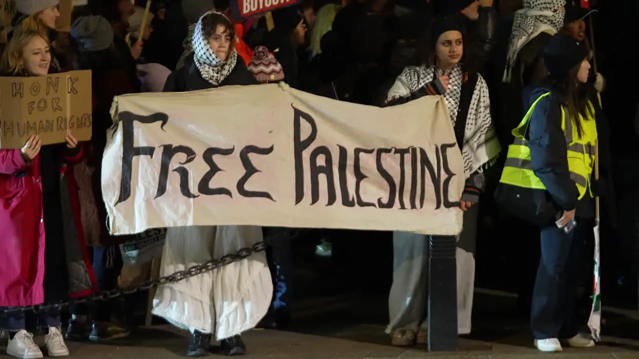 Two women wearing Keffiyeh scarves hold a white banner that reads Free Palestine” during a nighttime protest outside the Houses of Parliament