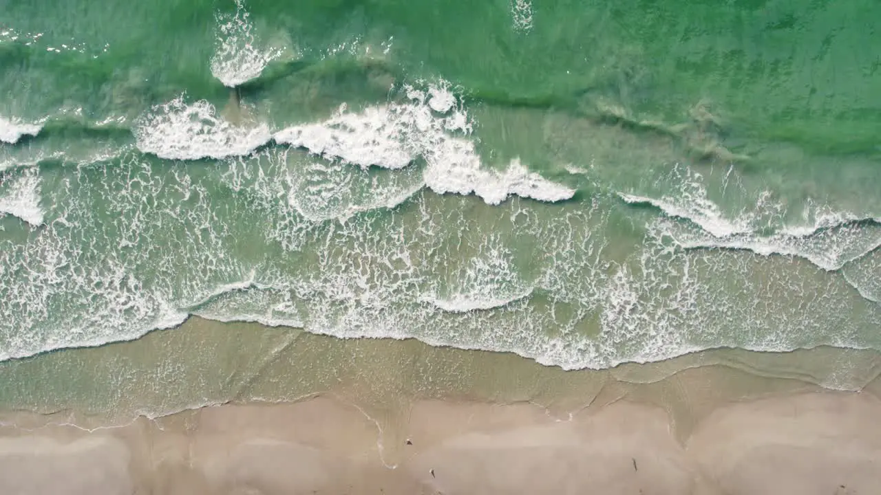 Aquamarine waves crash against warm golden sand from a bird's eye view in a drone in slow motion