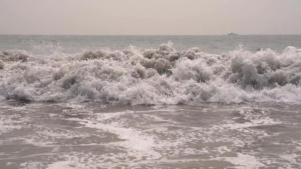 Close up of Pacific ocean waves crashing down onto the sandy beach in slow motion in San Diego California with a large ship in the background