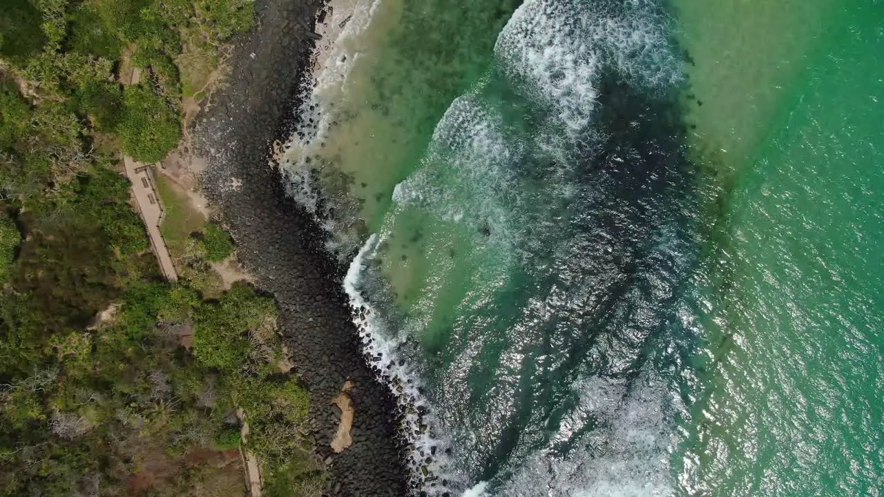 Looking vertically at small waves breaking on rocky beach adjacent walking track bright summers day drone