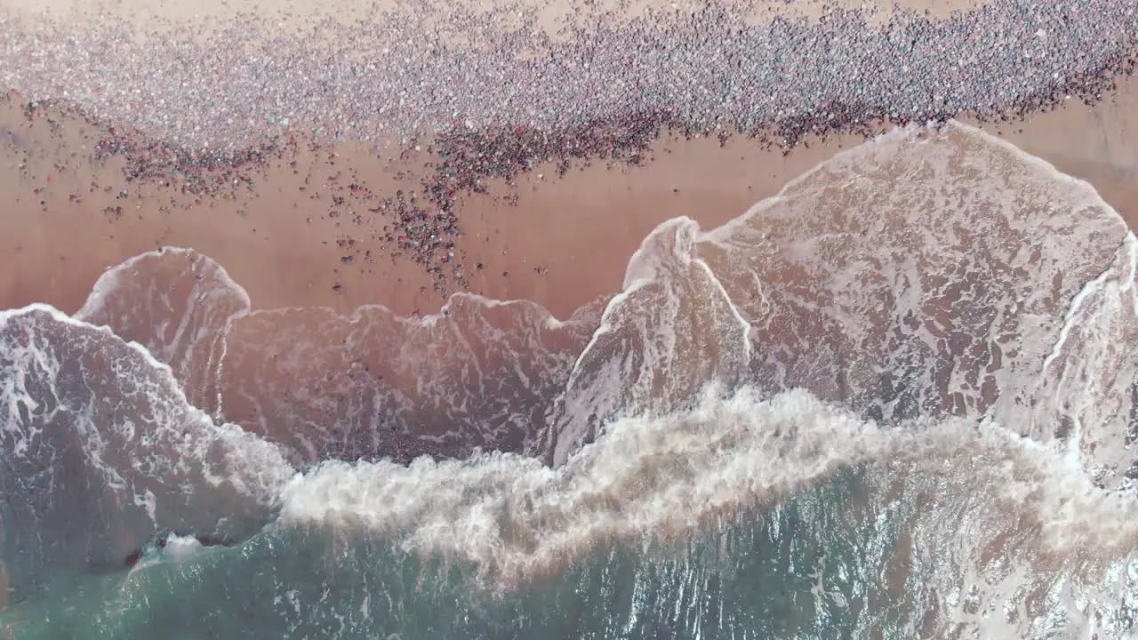 High aerial looking down at waves crashing on an abandoned pinkish red beach