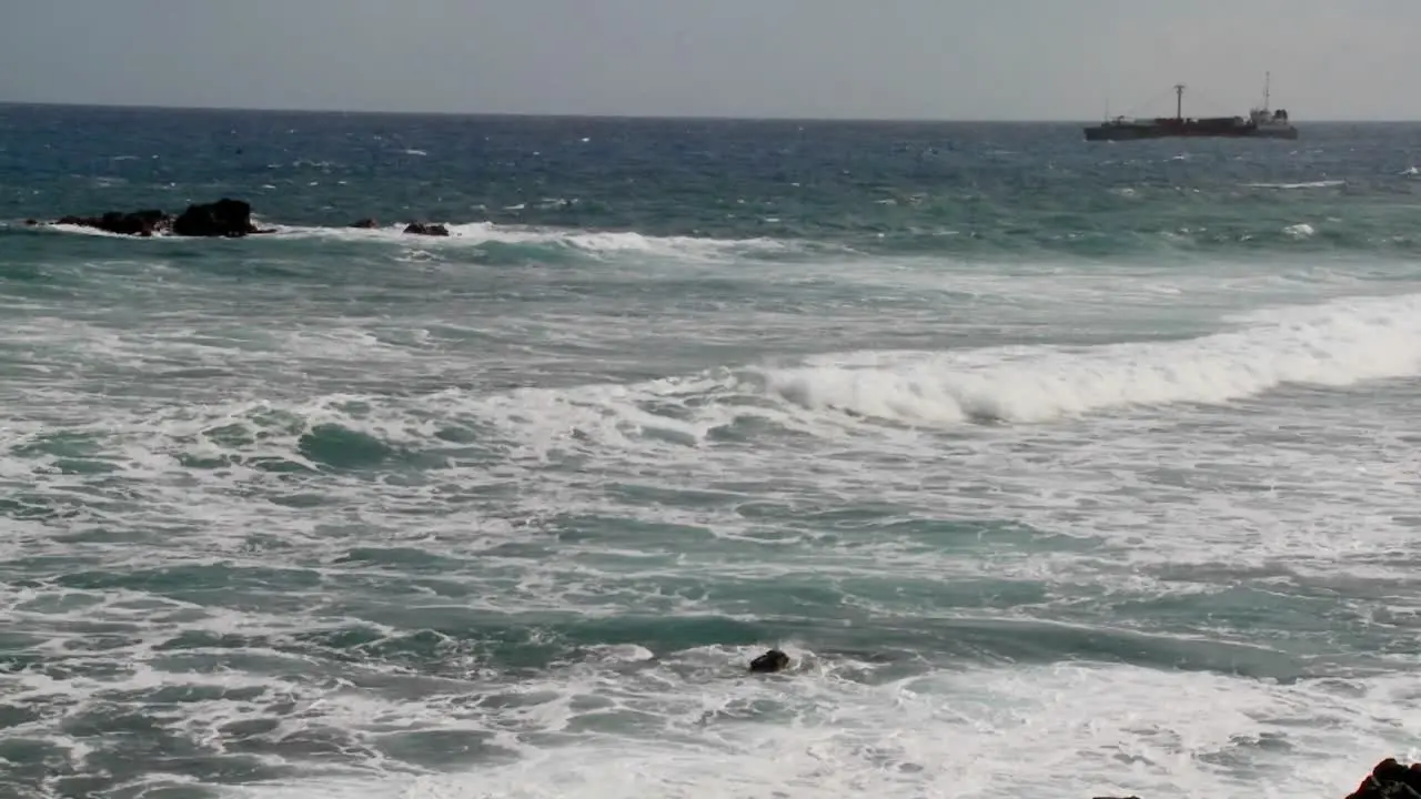 Waves roll into a rocky shore with a freighter ship in the background