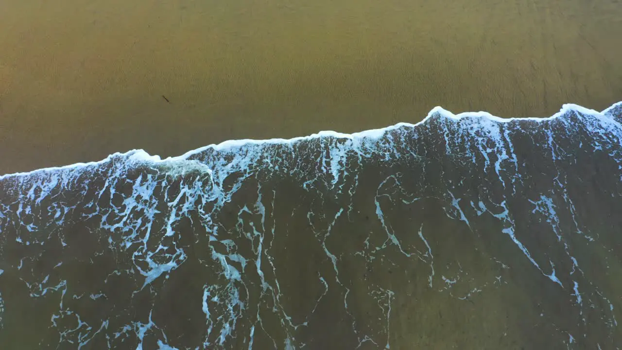 A drone captures a dramatic view of the ocean's energy as waves crash onto the sandy shore creating frothy white foam