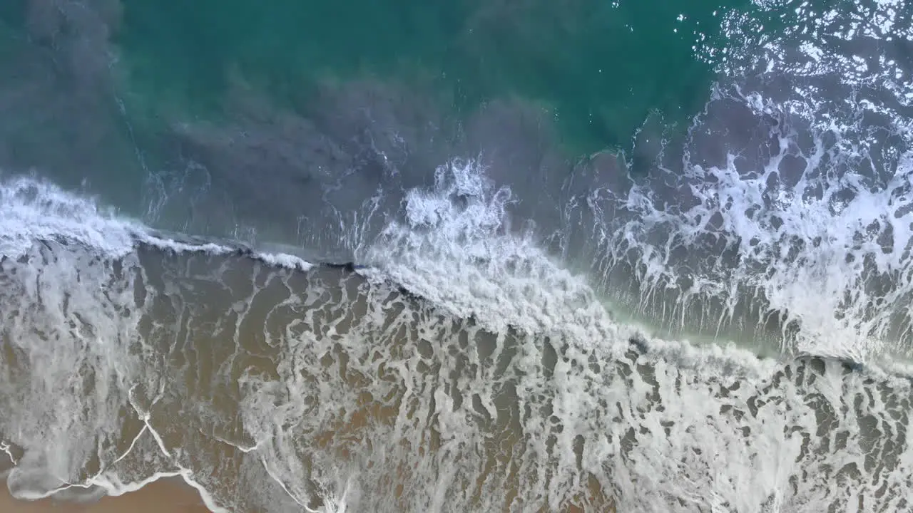 Aerial top-down shot overhead flying along a beach with the waves rolling in during the afternoon