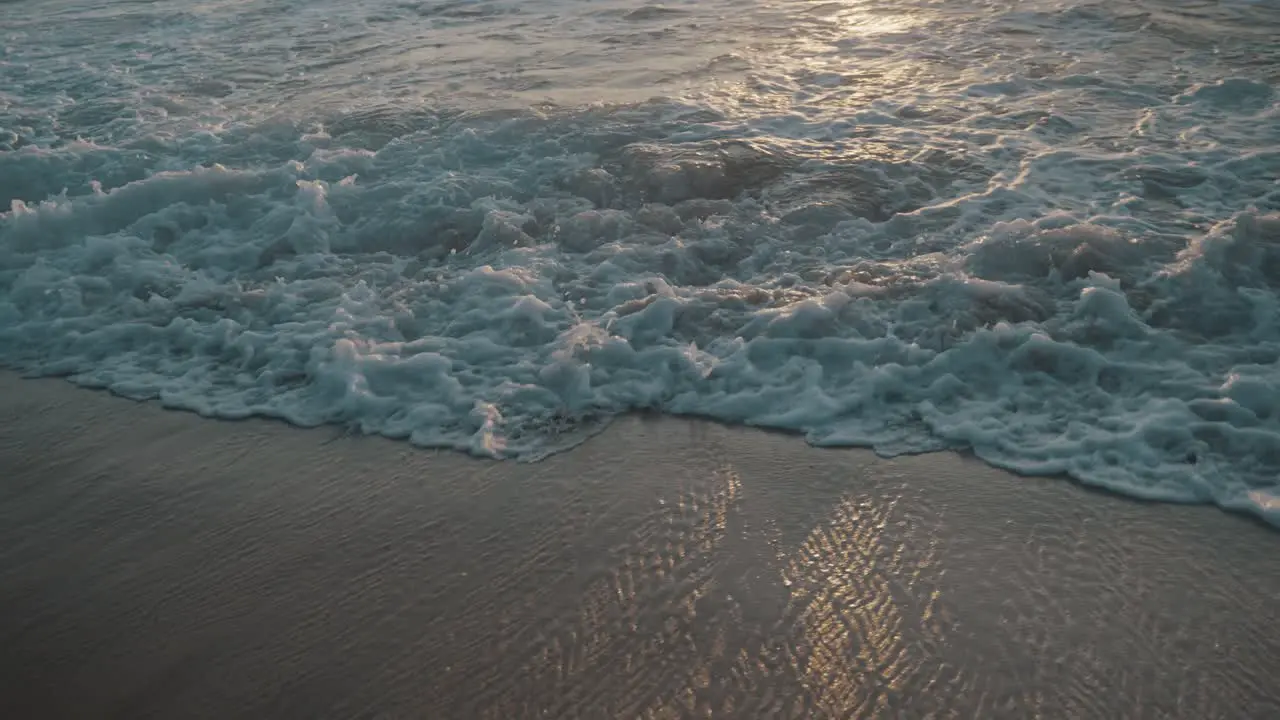 Water receding as the next wave rolls out onto the brown beach with a pile of white foam as the setting sun is reflected