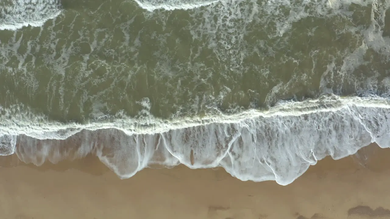 Aerial shot of crashing waves and a man with elongated shadow standing at the edge of the waves