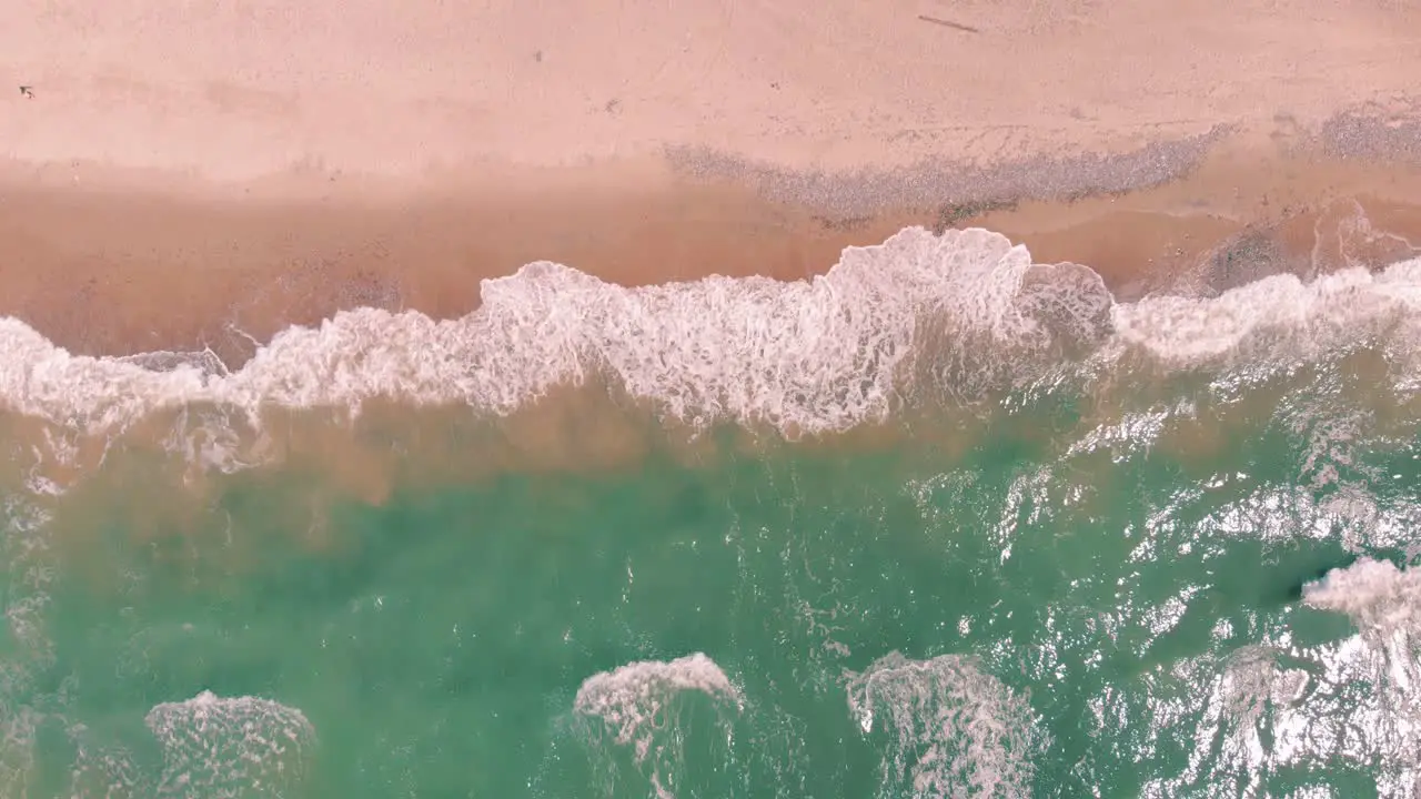 Static aerial shot of breathtaking sea-green waves crashing against pink sandy beach