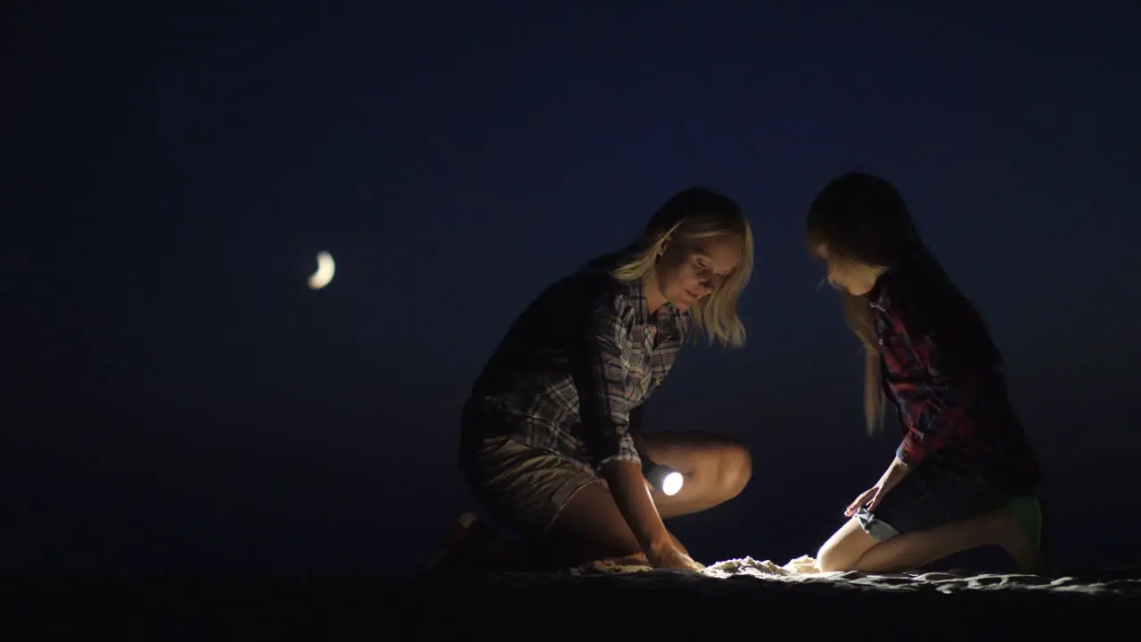 Mom And Daughter Are Playing Together In The Sand At Night They Shine With A Flashlight Looking For 