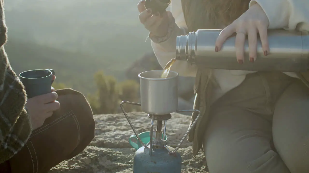 Close Up Shot Of An Unrecognizable Girl Pouring Hot Tea From Thermos Into Mug While Sitting At Mountain Top And Enjoying Hiking Trip With Friend