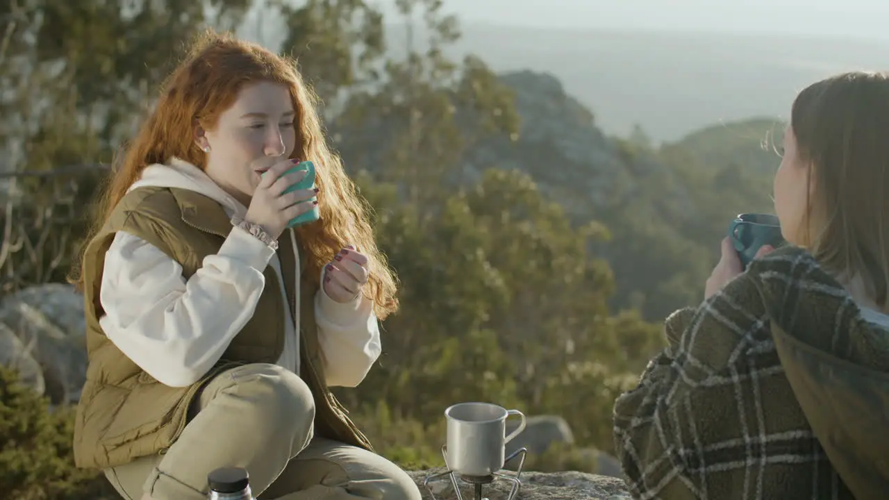 Two Young Female Hikers Drinking Hot Tea From Thermos While Sitting At Mountain Cliff On A Sunny Autumn Day 1