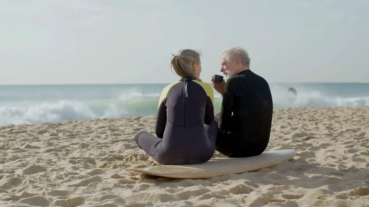 Long Shot Of A Senior Couple In Wetsuit Drinking Tea On Beach After Surf Training