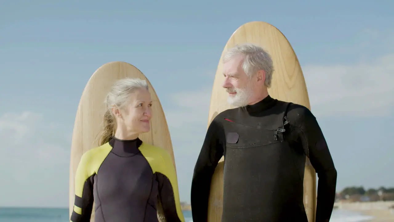 Front View Of A Senior Couple With Surfboard Behind Their Backs Standing On The Ocean Shore And Looking Ath The Camera