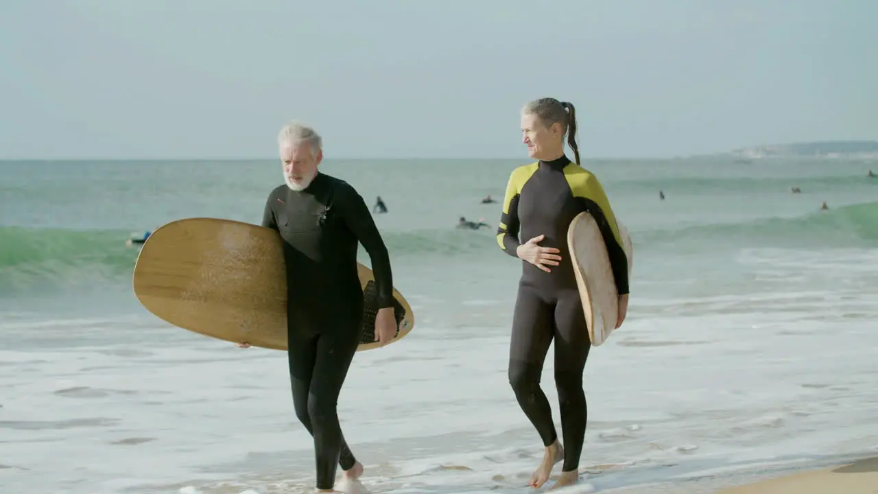 Senior Couple In Wetsuit Holding Hands And Walking Along The Beach With Surfboard