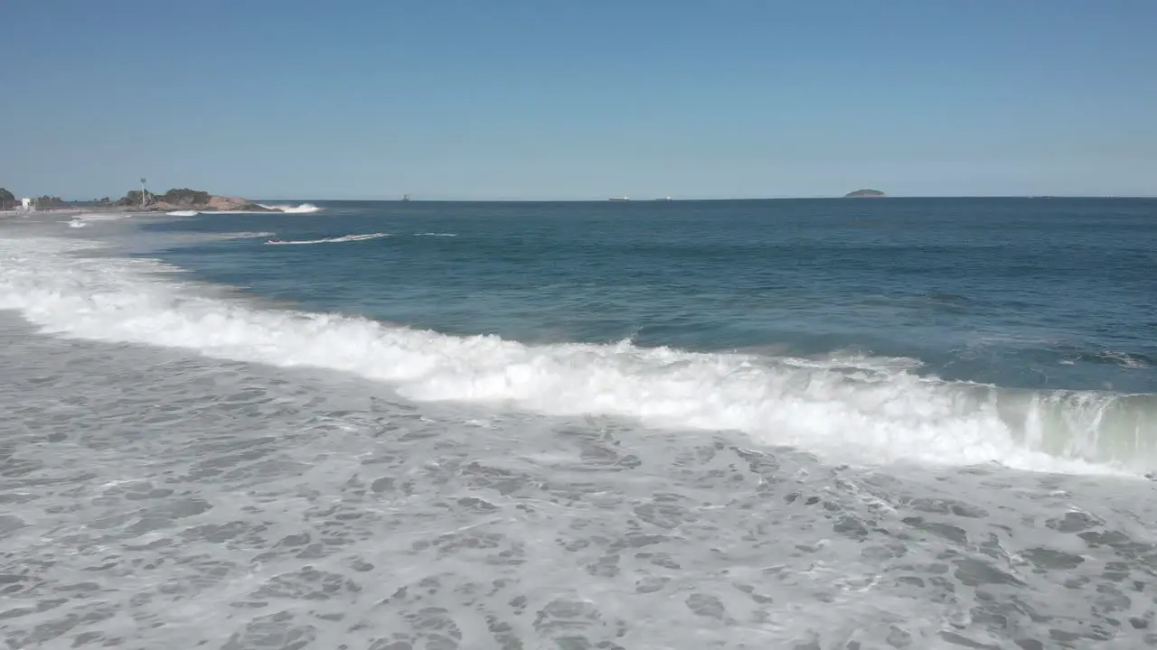 Backwards aerial movement following the incoming rough long waves coming in on Ipanema beach with the Arpoador rock and islands in the background on a sunny day in Rio de Janeiro