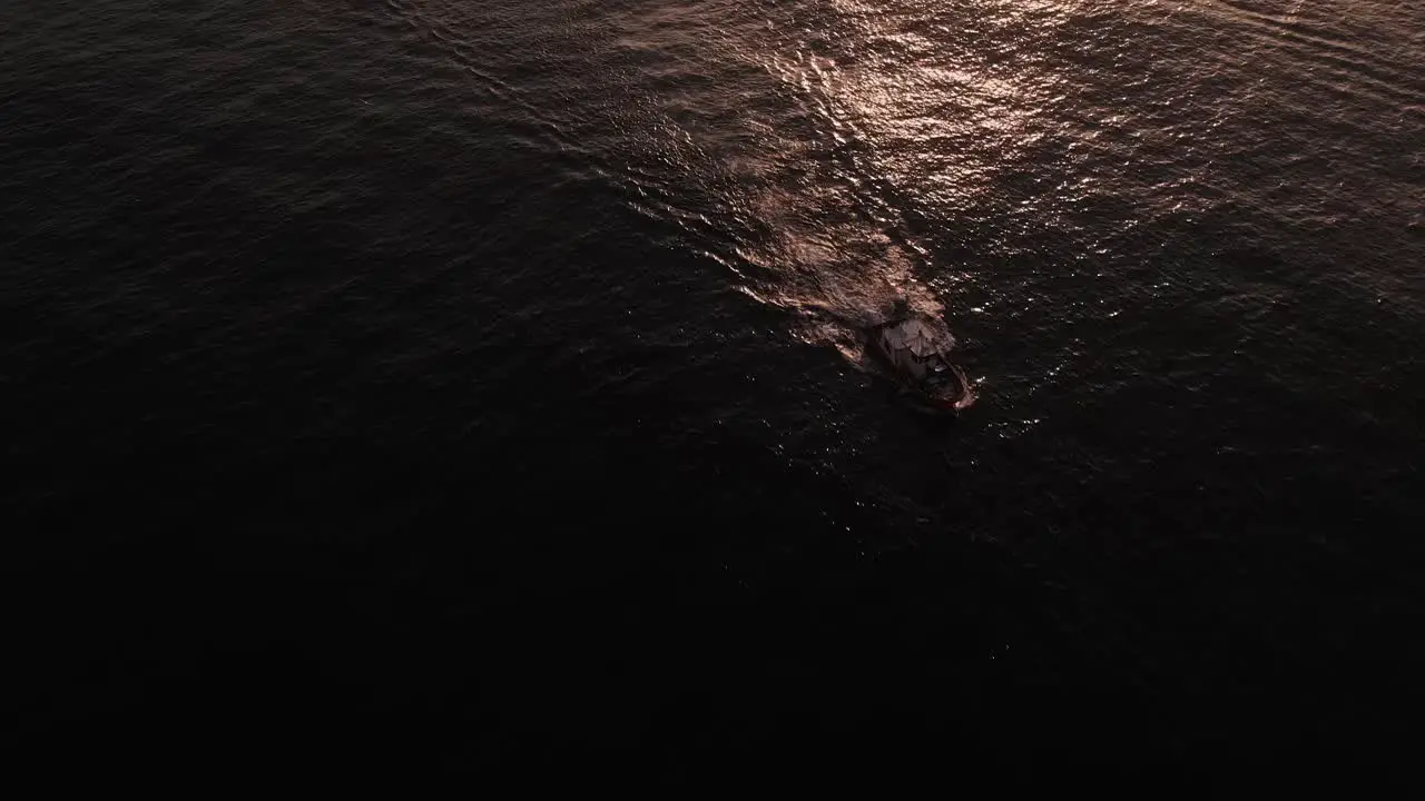 Aerial overlooking silhouetted boat travelling on quiet shimmering ocean waves tilt up to sunset skyline