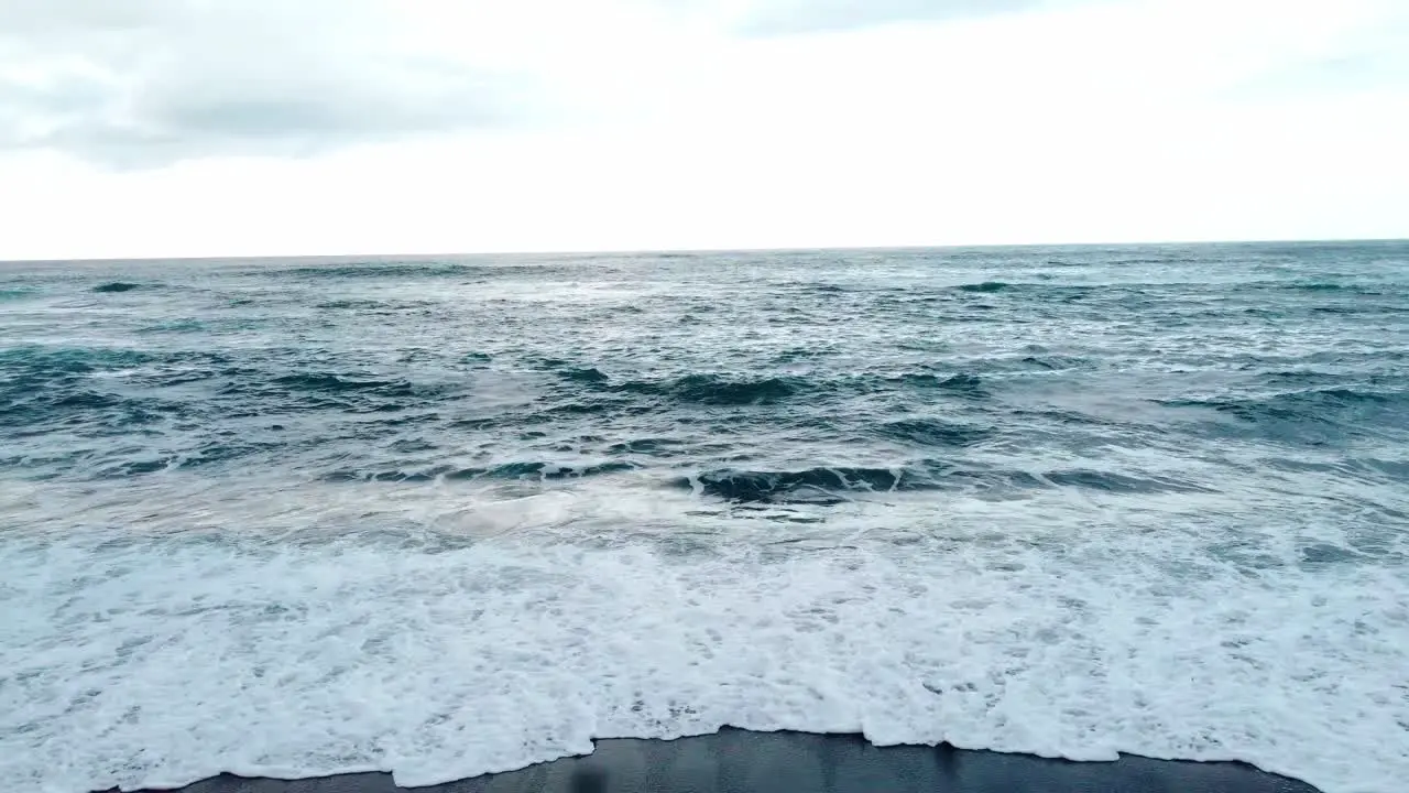 Pull drone shot of a blonde girl walking in a black sand beach in canary island with rough ocean in a cloudy moody day