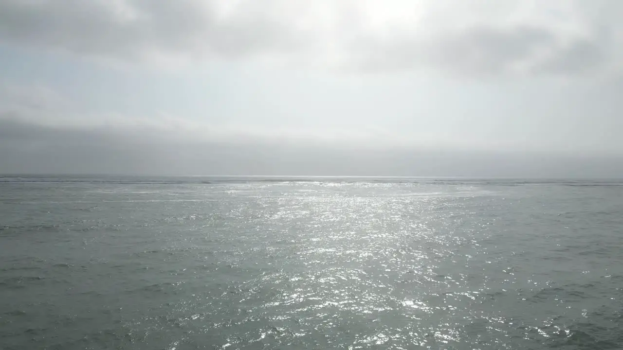 Low angle aerial drone shot over the sea on a sunny day reflecting the sun on the water with view of the horizon with surfern with surfboards in water