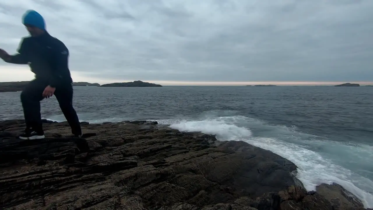 Male adult sitting on rock close to sea and using his cellphone before raising up and walking away Static clip with waves washing up at shore and dramatic clouds after sunset