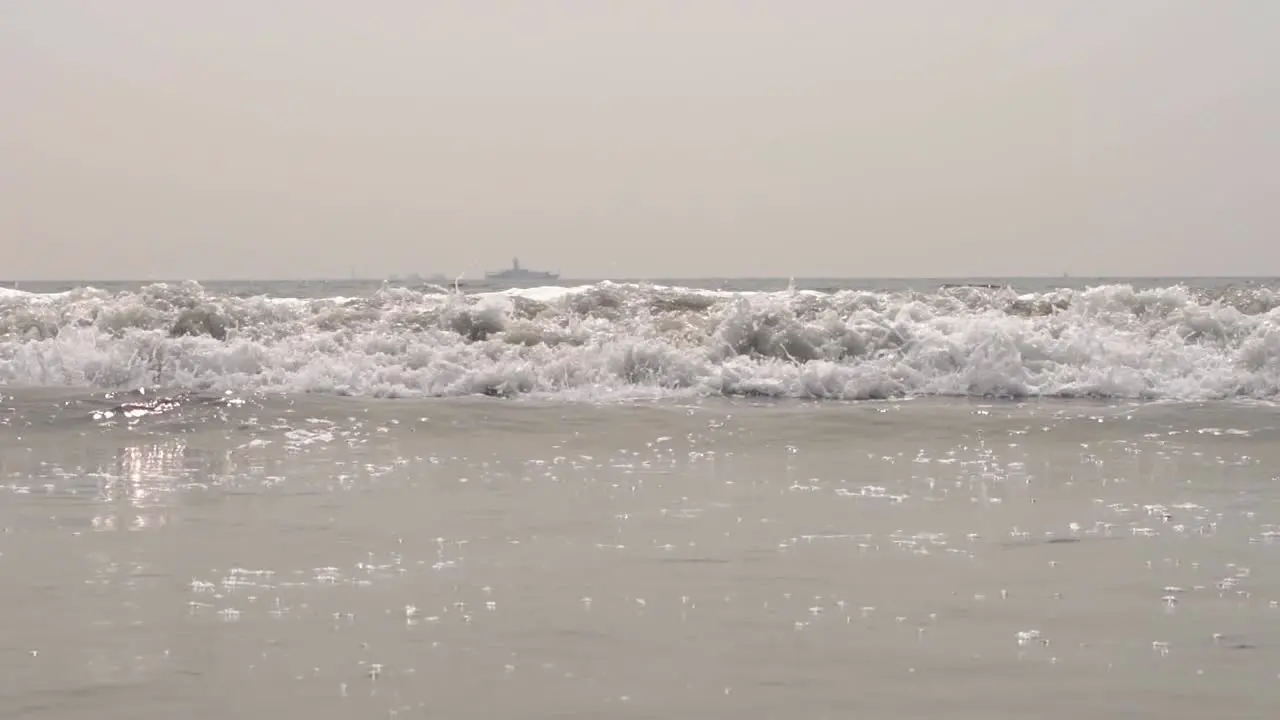 Pacific ocean waves crashing down onto the sandy beach in slow motion in San Diego California with a large ship in the background wide shot