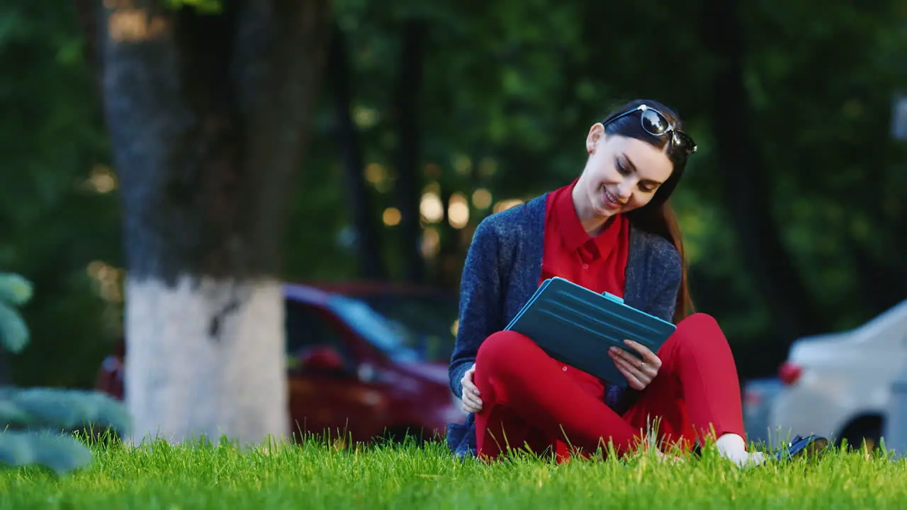 Stylish Woman Sitting On The Grass In The Park Enjoying The Tablet In The Background People Walk